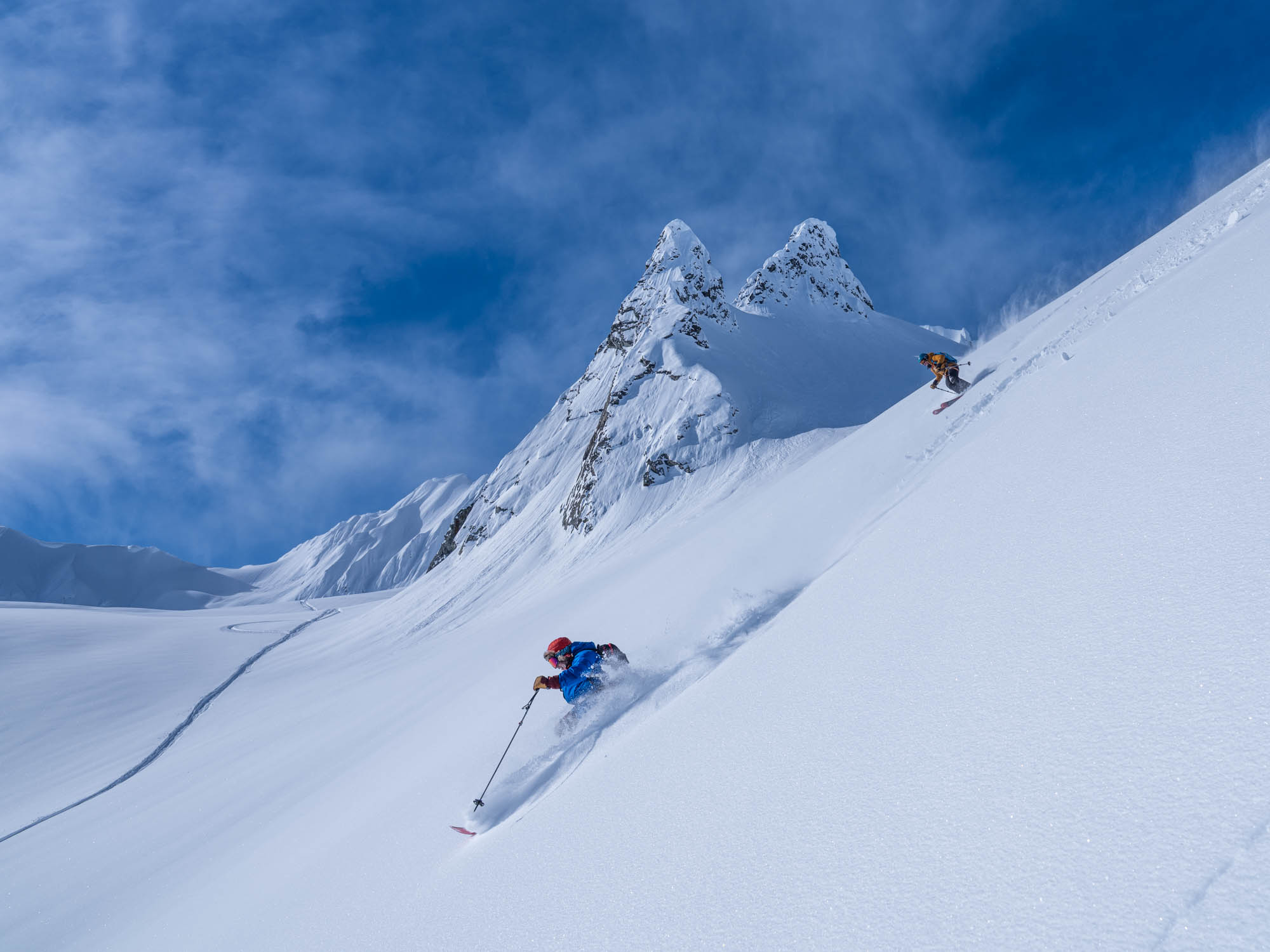 Sean and Rihianna Brownell carve fresh lines down a pristine, powder-filled slope in Haines, Alaska, with jagged peaks rising in the background — showcasing the epic terrain and pure joy of heli-skiing with Alaska Heli Skiing.
